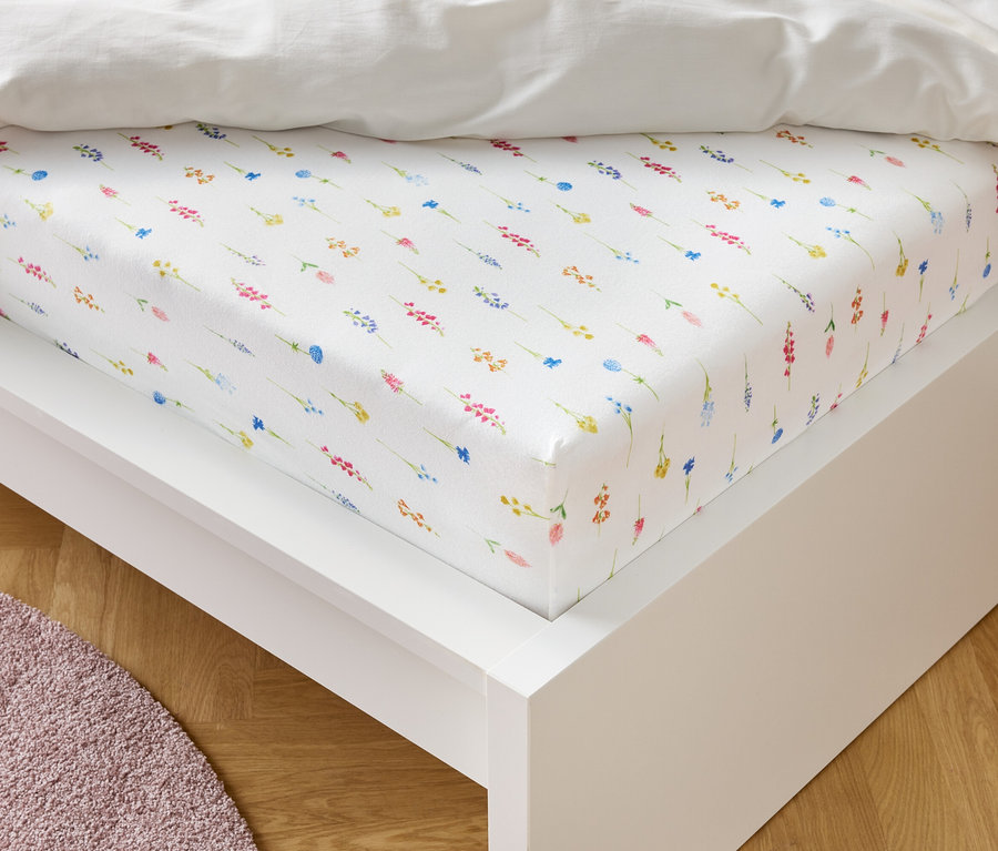 Biele napínacie prestieradlo s kvetinovým vzorom leží na bielom ráme postele.