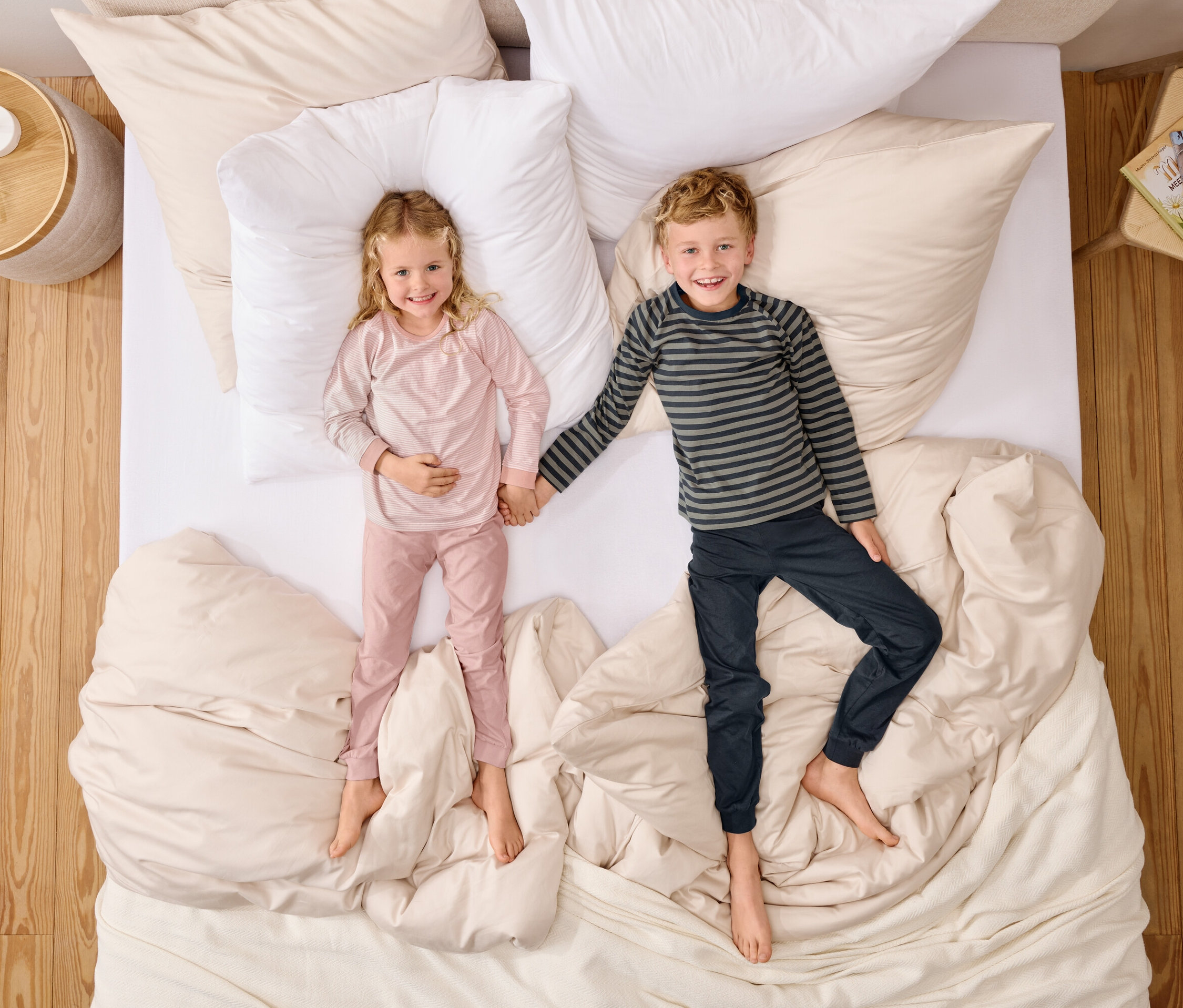 Pohľad zhora na dievča v ružovom pyžame a chlapca v pruhovanom tope, ktorí sa držia za ruky na posteli s bielymi vankúšmi.