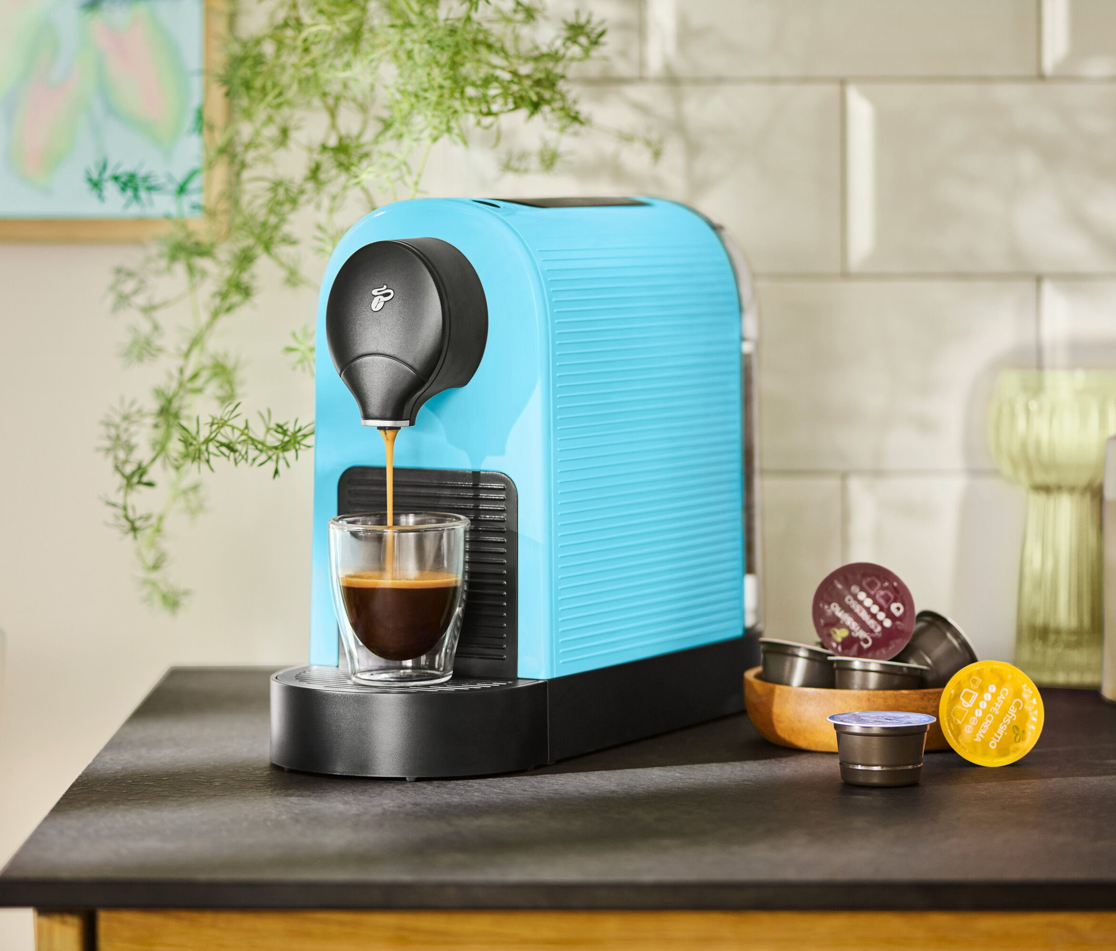 Tyrkysový kávovar plní šálku kávou. Vedľa v drevenej miske ležia kávové kapsuly.