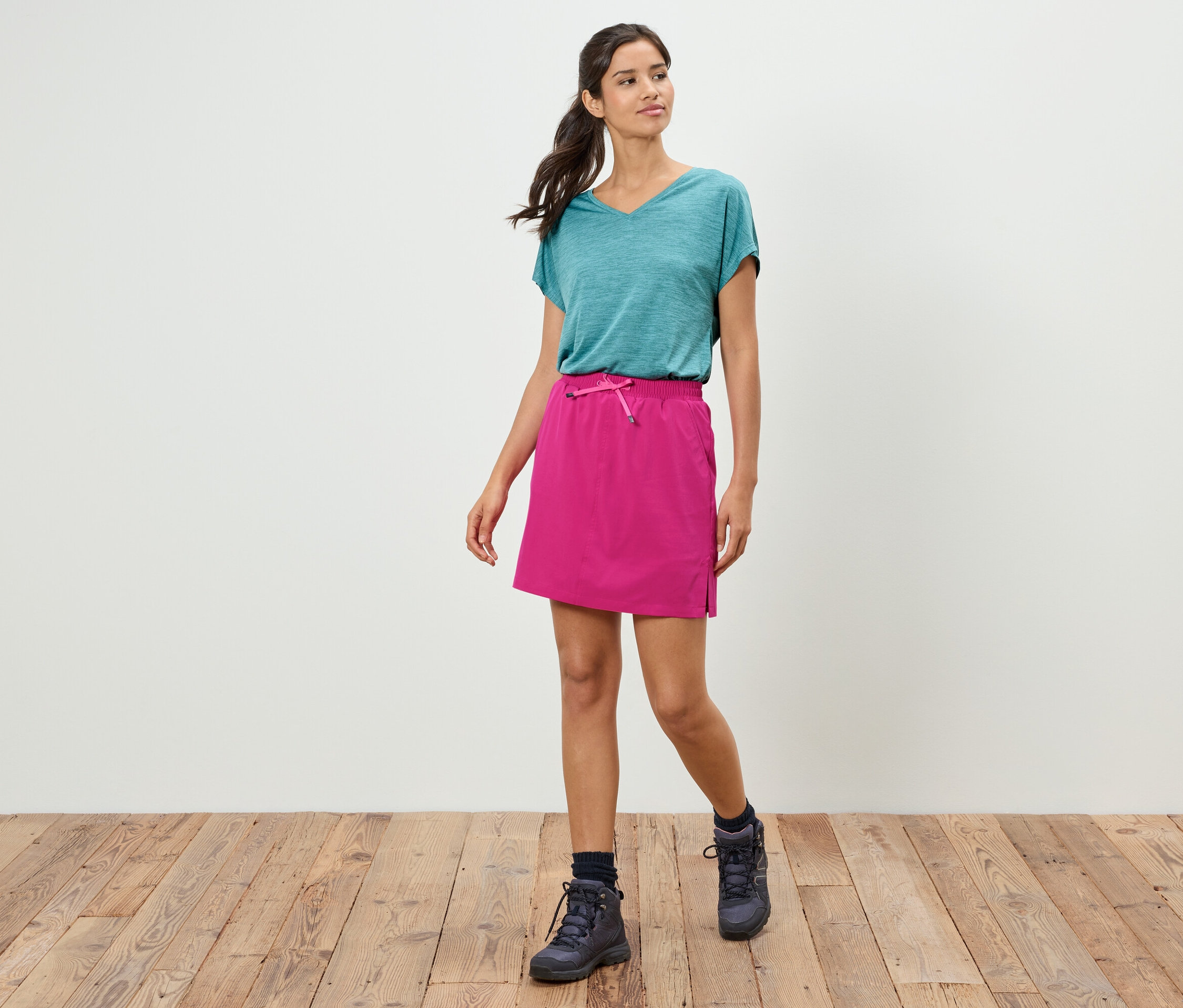 Žena kráča v tyrkysovom tričku, ružovej sukni a trekových topánkach po drevenej podlahe pred bielou stenou.