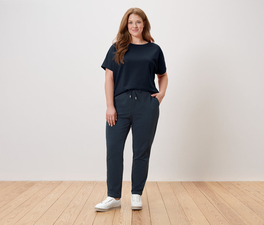 Žena stojí pred stenou a má na sebe tmavomodré tričko, tmavomodré nohavice a biele topánky.