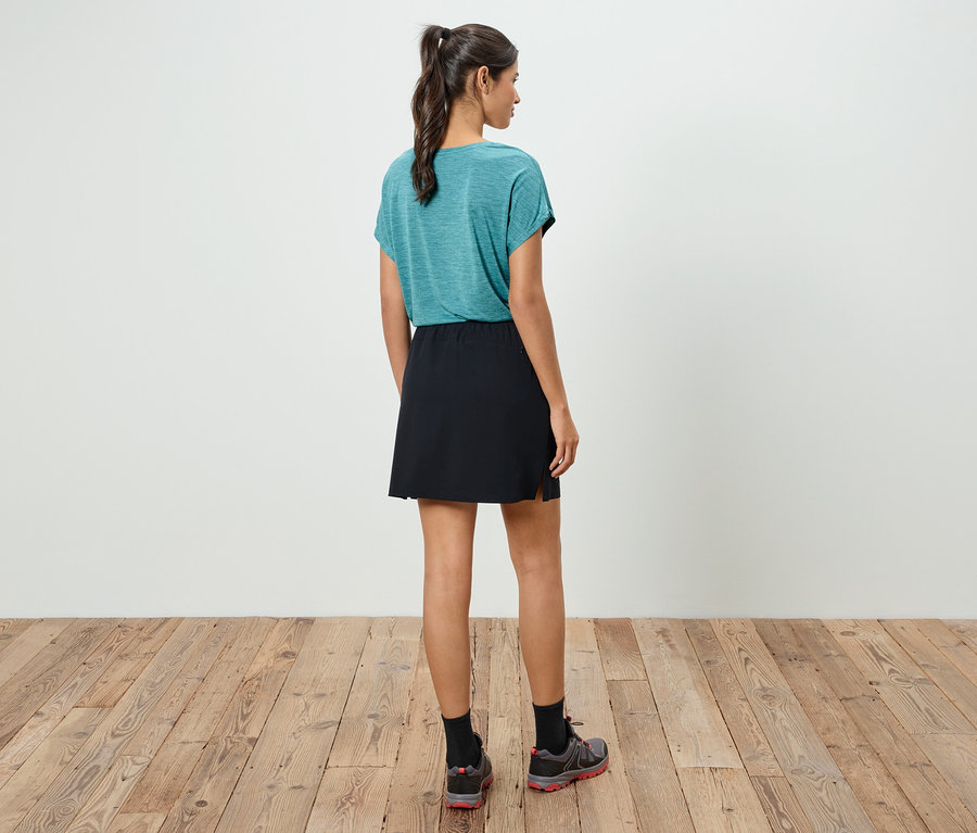 Žena stojí chrbtom k fotoaparátu, má na sebe modrozelené tričko, čiernu sukňu a športové topánky.