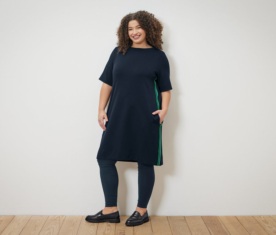Žena s kučeravými vlasmi má na sebe tmavomodré šaty so zelenými pruhmi, tmavomodré legíny a čierne mokasíny.