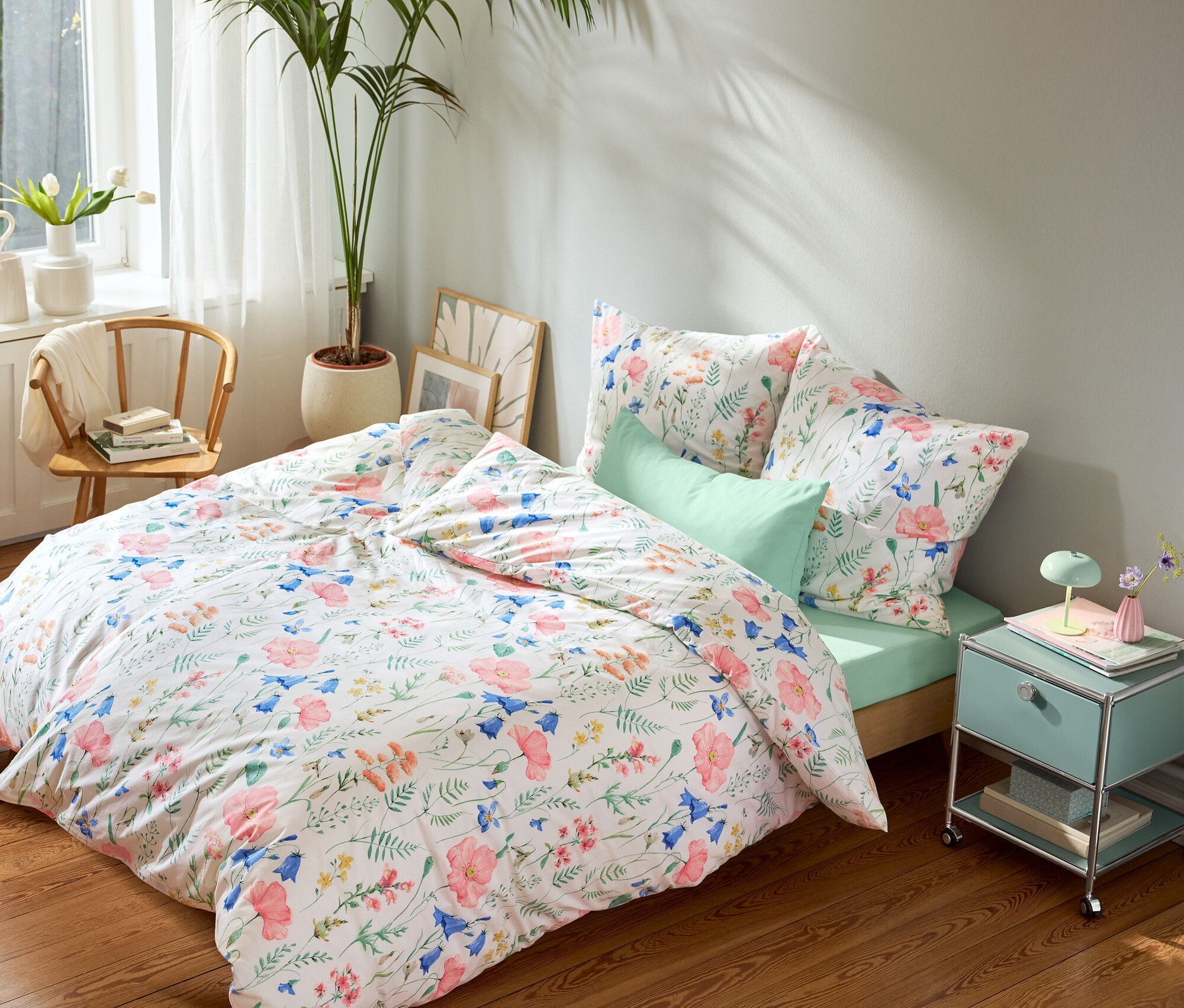 Posteľ s kvetinovou posteľnou bielizňou, mätovo zelenou plachtou a mätovo zelenými obliečkami na vankúše, mätovo zelený nočný stolík a okrúhly koberec.