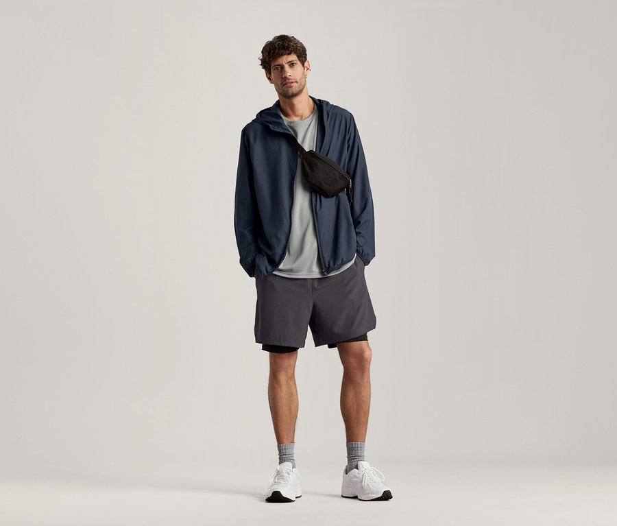 Muž stojí a má na sebe svetlosivé športové tričko s melírom, tmavomodrú bundu a sivé šortky.