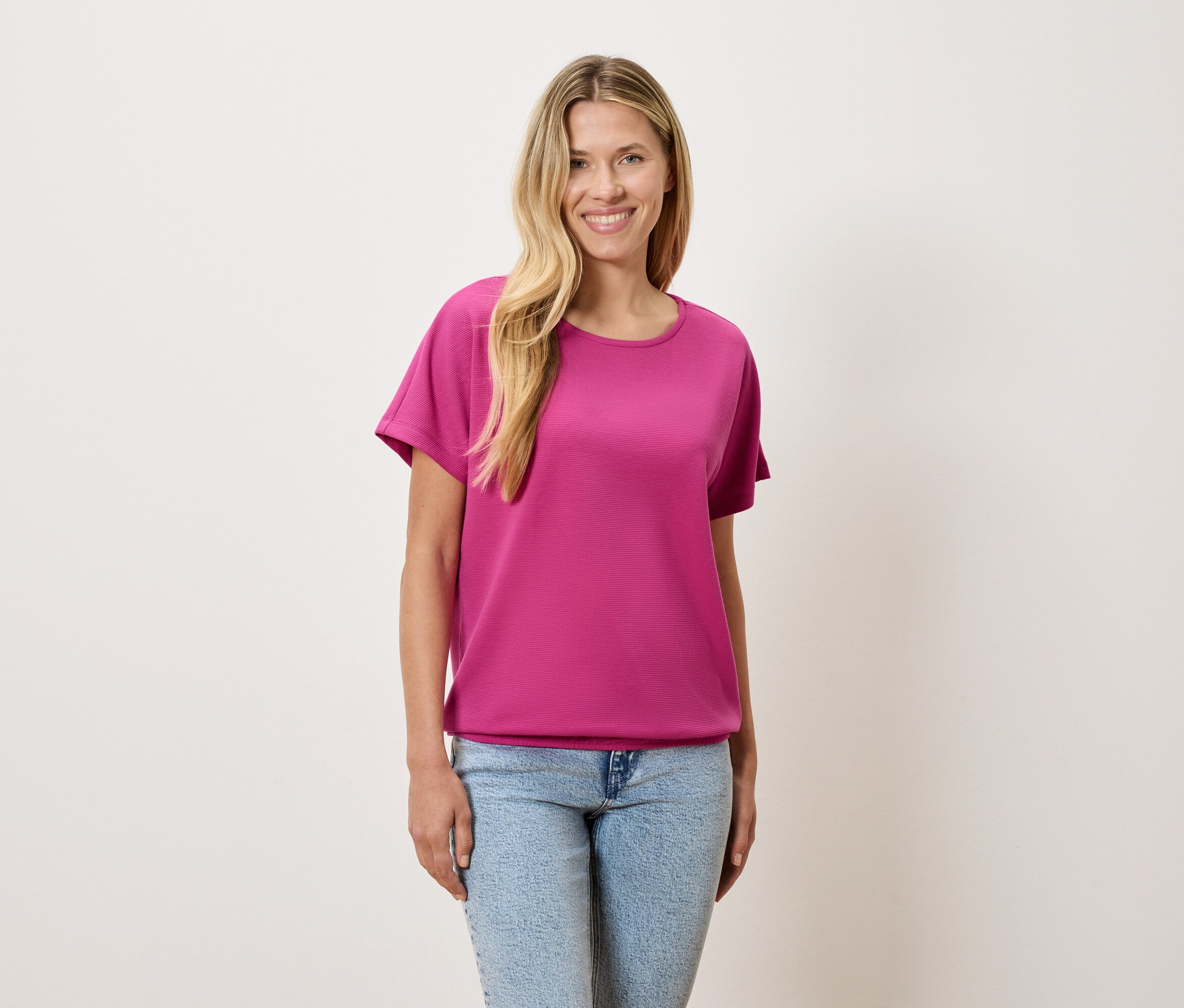 Žena s blond vlasmi má na sebe ružové tričko a modré rifle.