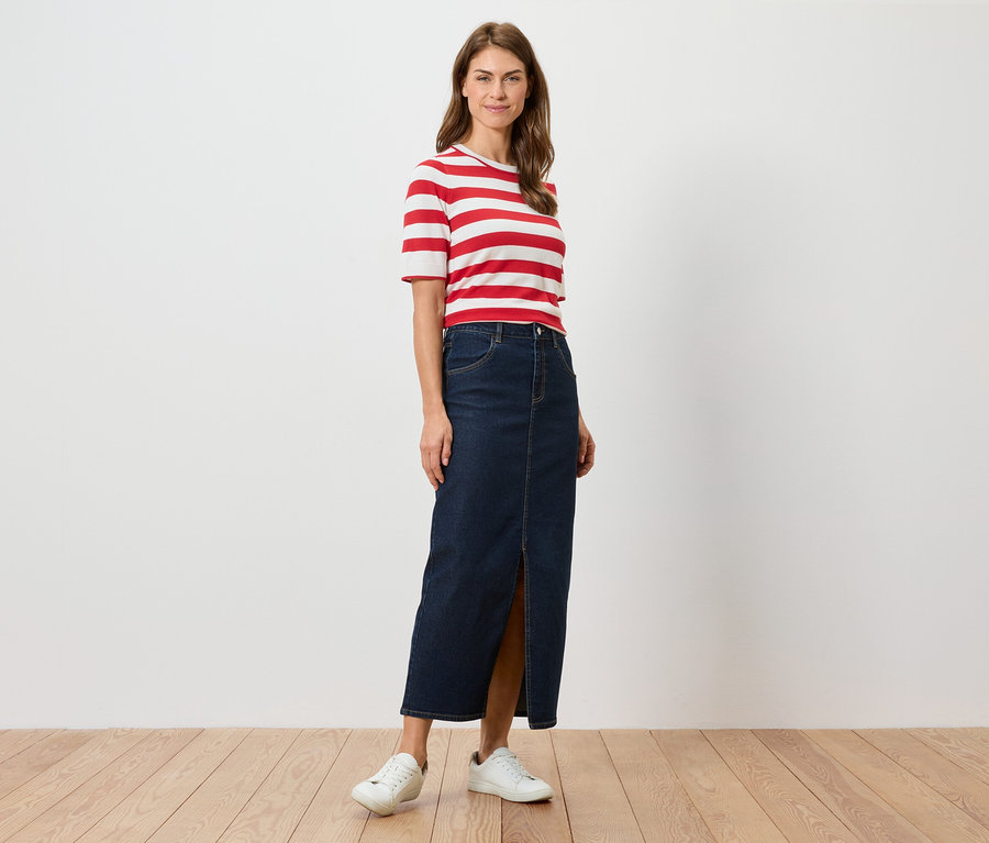 Žena stojí a má na sebe červeno-biele pruhované tričko, dlhú džínsovú sukňu s rozparkom a biele tenisky.