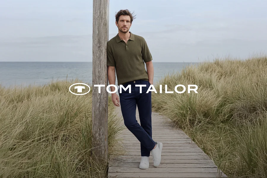 Muž v zelenom polo tričku, tmavomodrých nohaviciach a bielych teniskách sa opiera o drevený stĺp na drevenom chodníku pri mori, obklopený vysokou trávou. Logo TOM TAILOR.