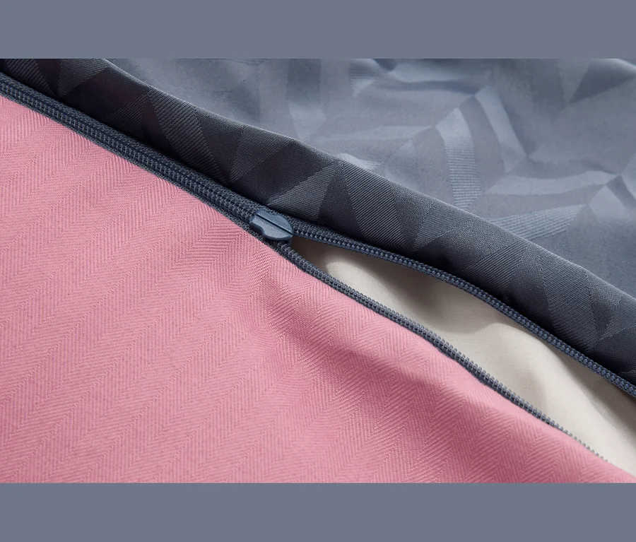 Detail tmavomodrého zipsu spájajúceho ružovú látku so vzorom rybej kosti a sivo-modrú látku s geometrickým vzorom, čiastočne otvorený, odhaľujúci svetlú látku pod ním.