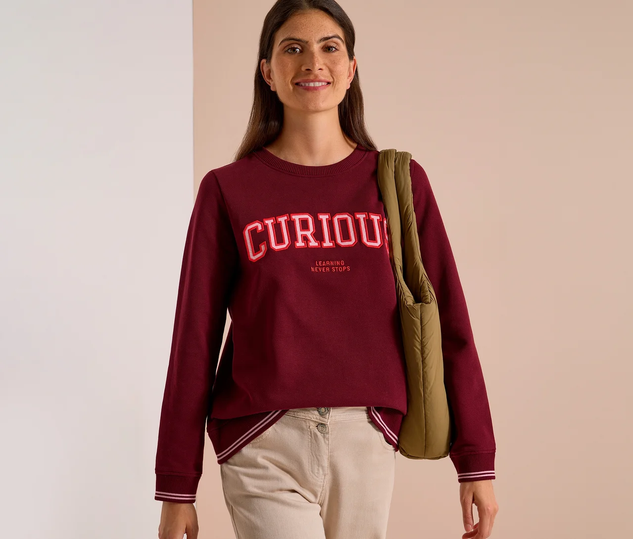 Usmievavá žena v bordovej Mikine CECIL s nápisom "CURIOUS LEARNING NEVER STOPS" a béžových nohaviciach, s prehodenou zelenou taškou cez rameno.