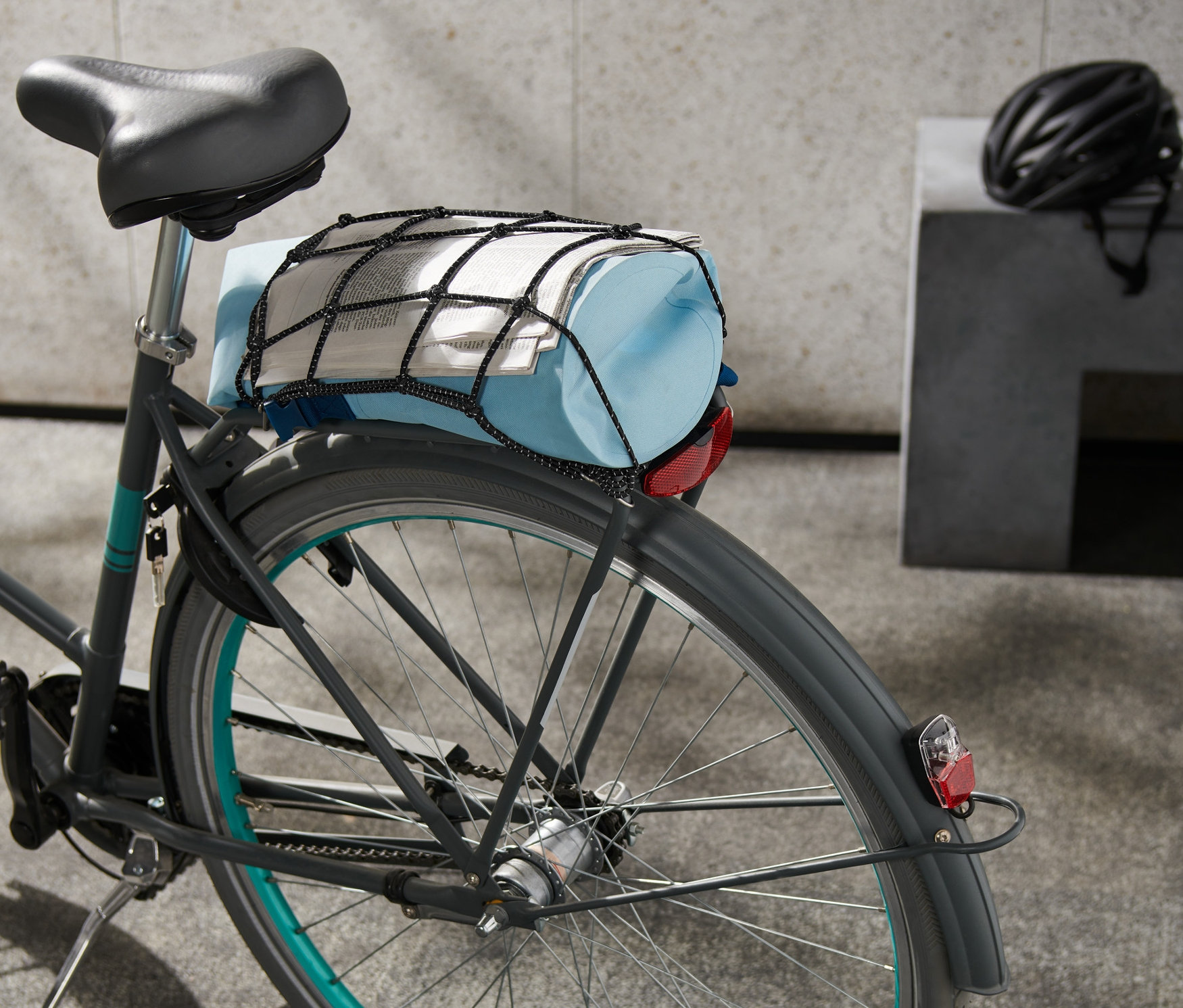 Na bicykli je sieť na batožinu na bicykel, ktorá drží modrú tašku a noviny.
