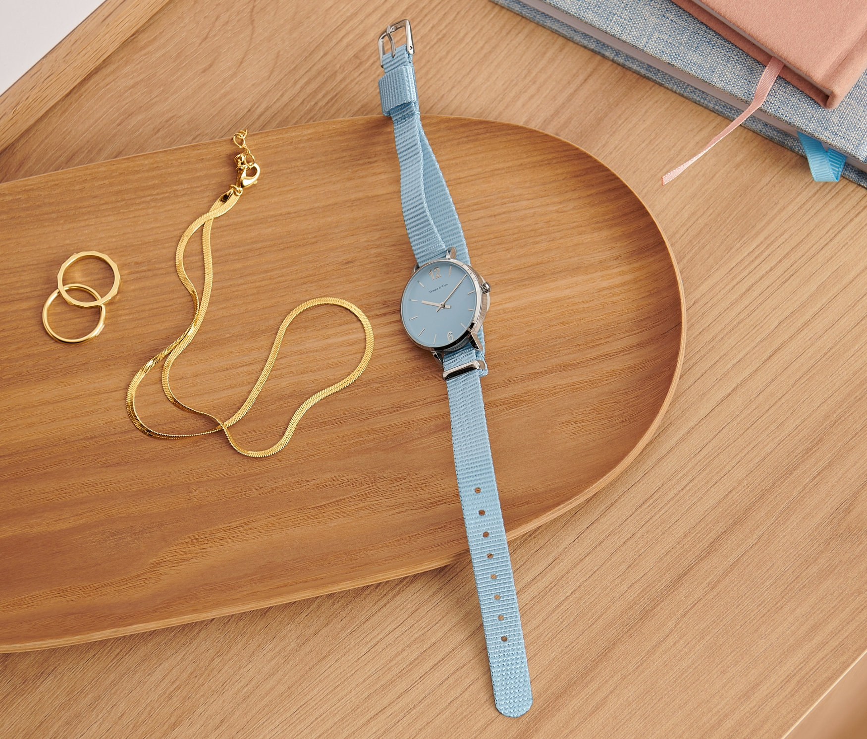 Modré quartzové náramkové hodinky z ušľachtilej ocele 316L ležia na drevenom podnose vedľa zlatého náhrdelníka a prsteňov.