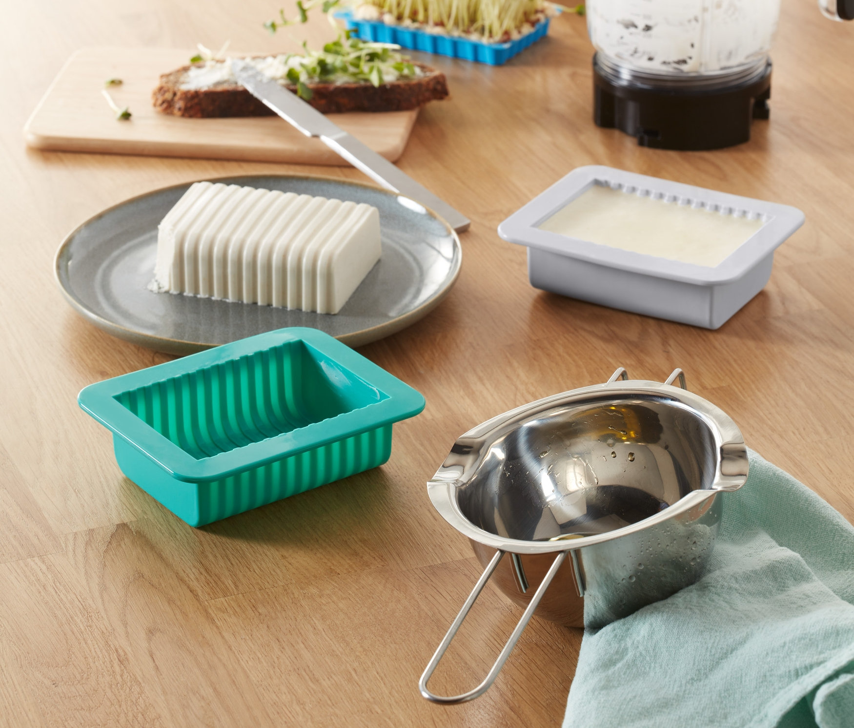 Na stole je súprava na vlastnoručnú výrobu rastlinného masla, forma na maslo, mixér, klíčky a chlieb s maslom.