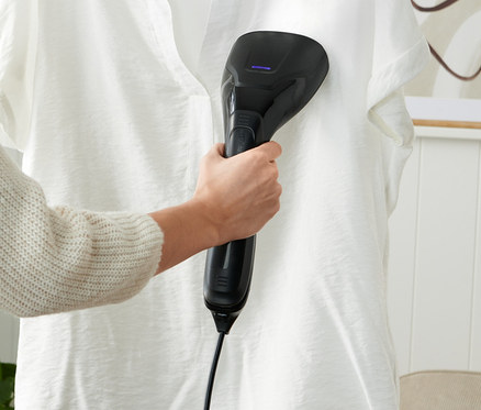 Žena používa naparovaciu kefu na bielu košeľu visiacu na vešiaku.