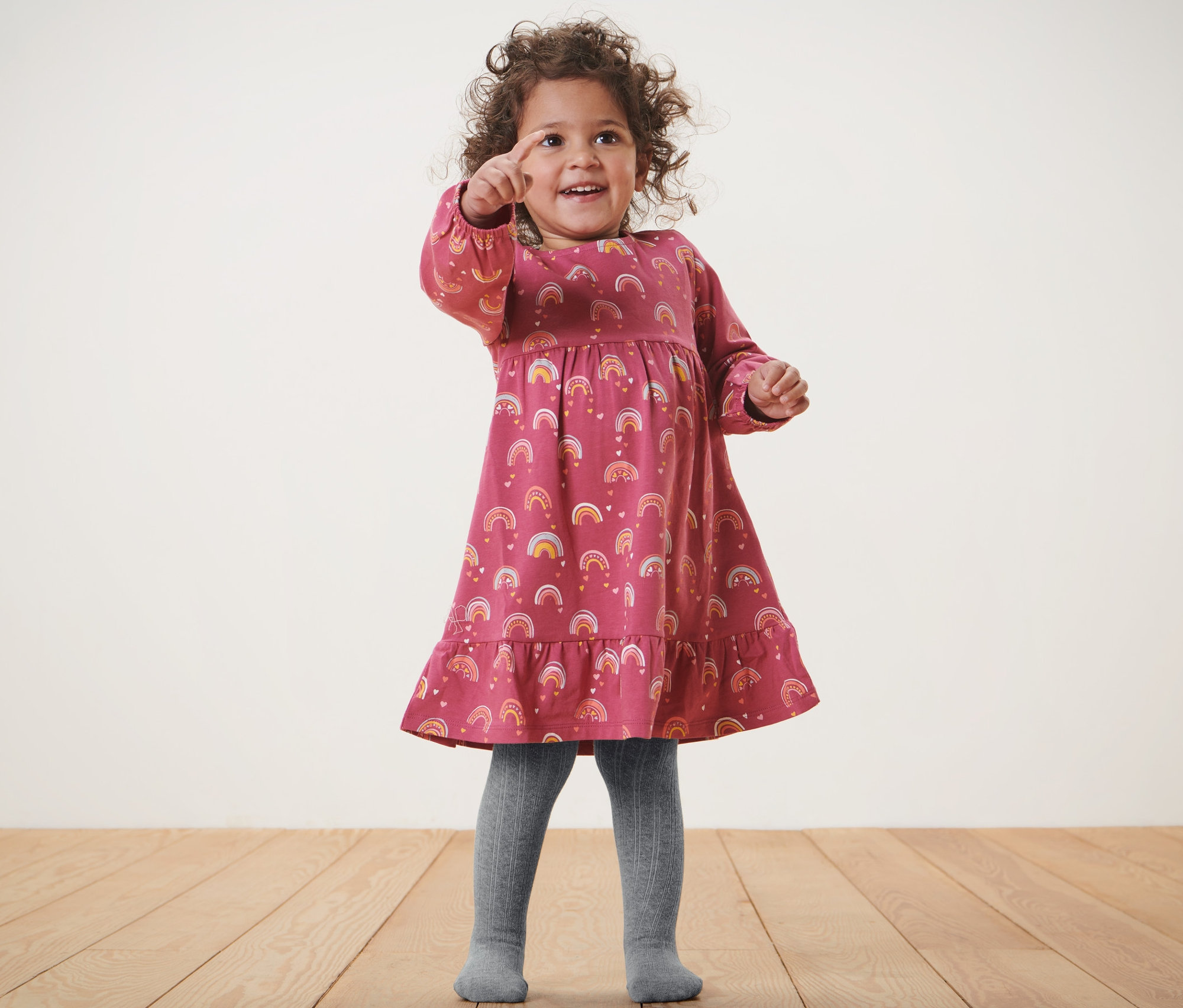 Dievča v šatách s dúhovou potlačou a sivých pančuchových nohaviciach pre bábätká, 2 páry, ukazuje prstom.