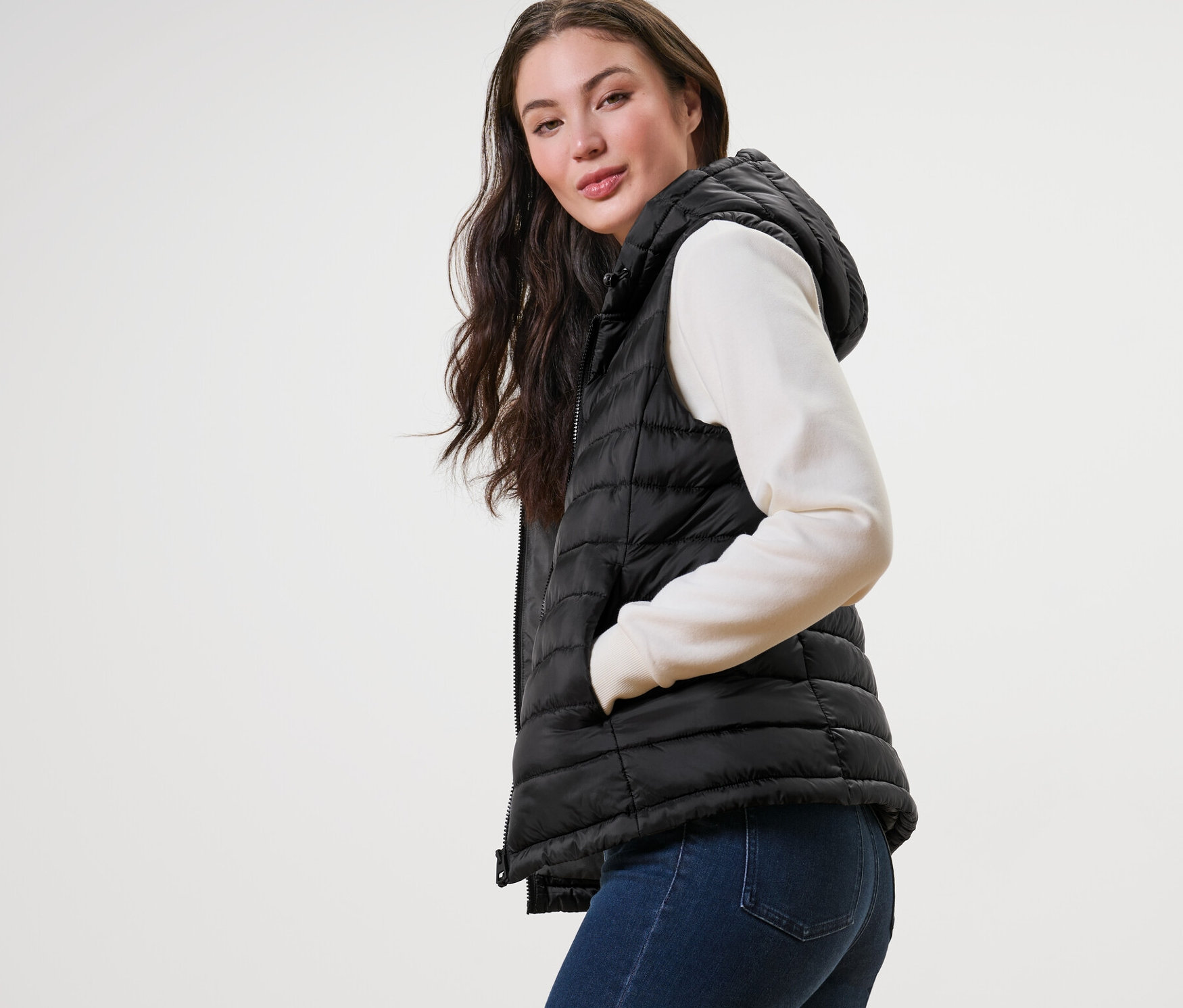 Žena pózuje v prešívanej veste s kapucňou, svetri a džínsoch.