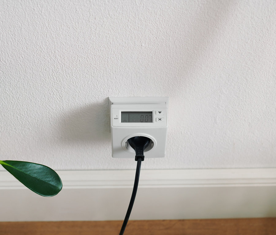 Biely merač spotreby elektrickej energie zapojený do bielej zásuvky na bielej stene s čiernym káblom.