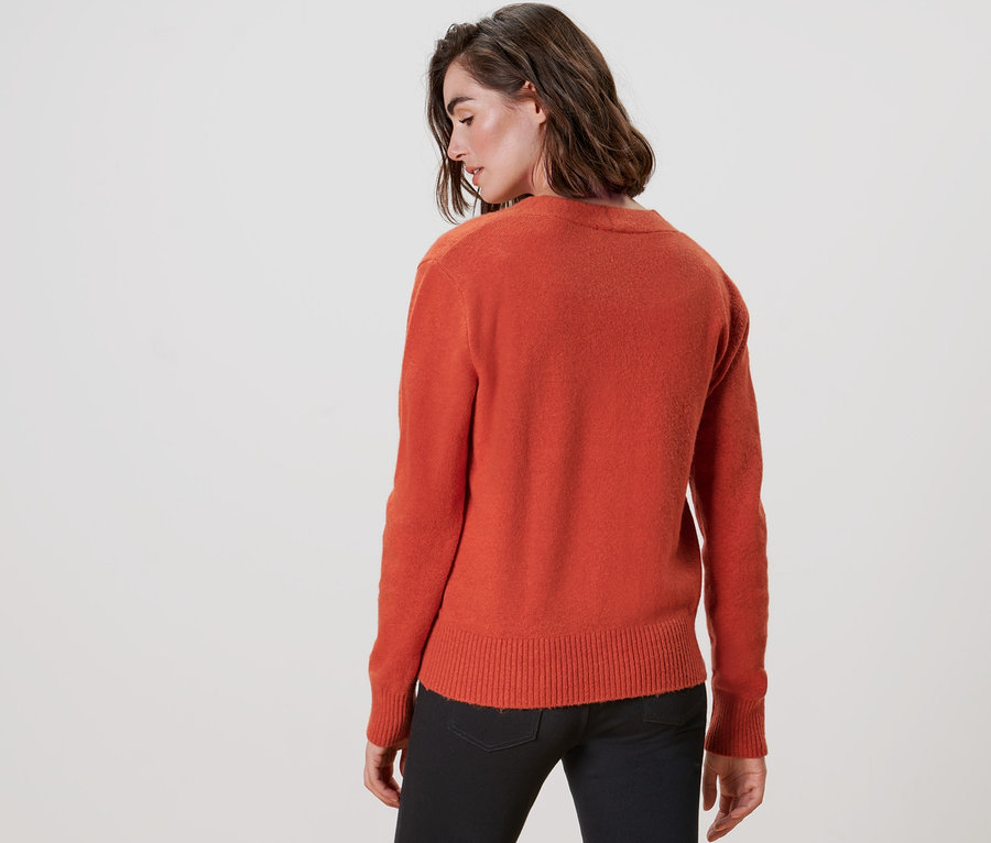 Zozadu je vidieť žena v oranžovom kardigáne s výstrihom do V a čiernych nohaviciach.