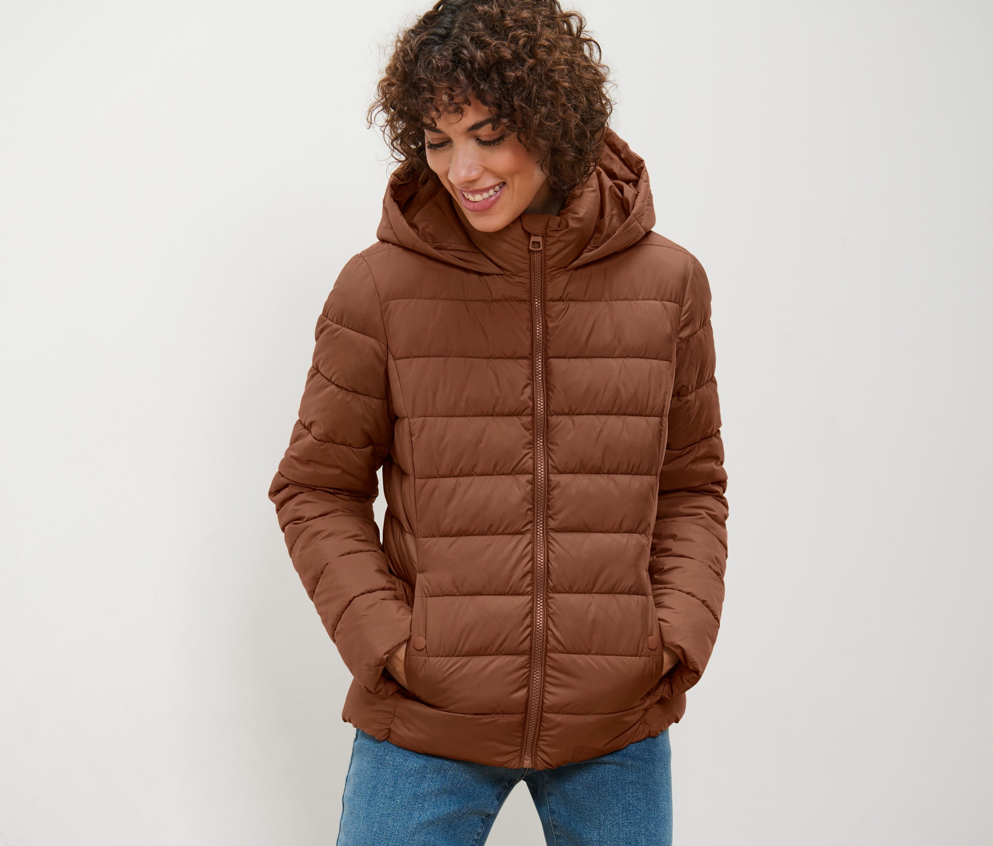 Žena má na sebe koňakovú prešívanú bundu s kapucňou a pozerá sa dole.
