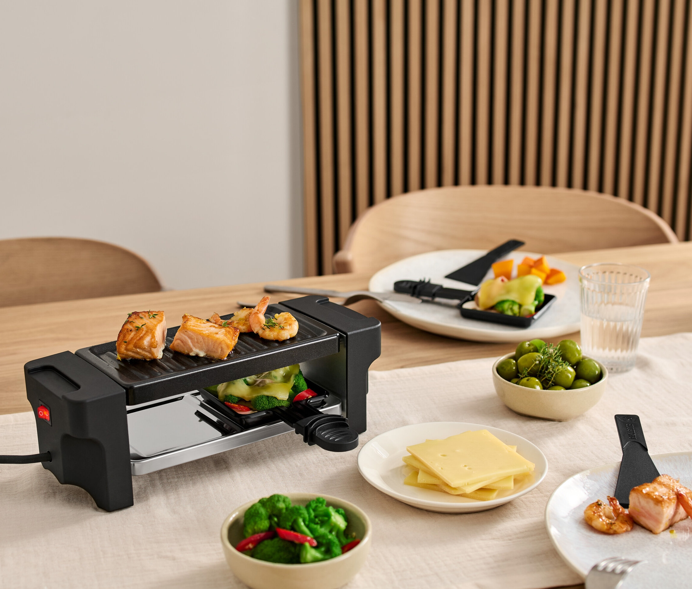 Na stole je zostava na raclette pre dvoch s lososom a krevetami na grile. Taniere s ingredienciami sú okolo grilu.