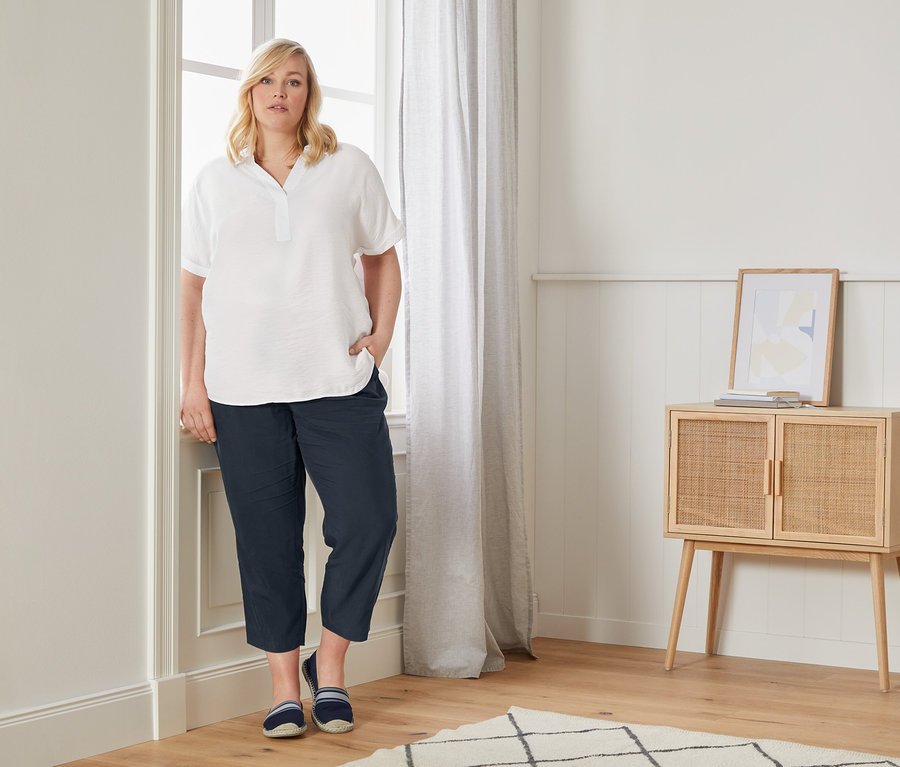 Žena pózuje pri okne v blúzke v oversize štýle, ležérnych nohaviciach chino a espadrilkách.