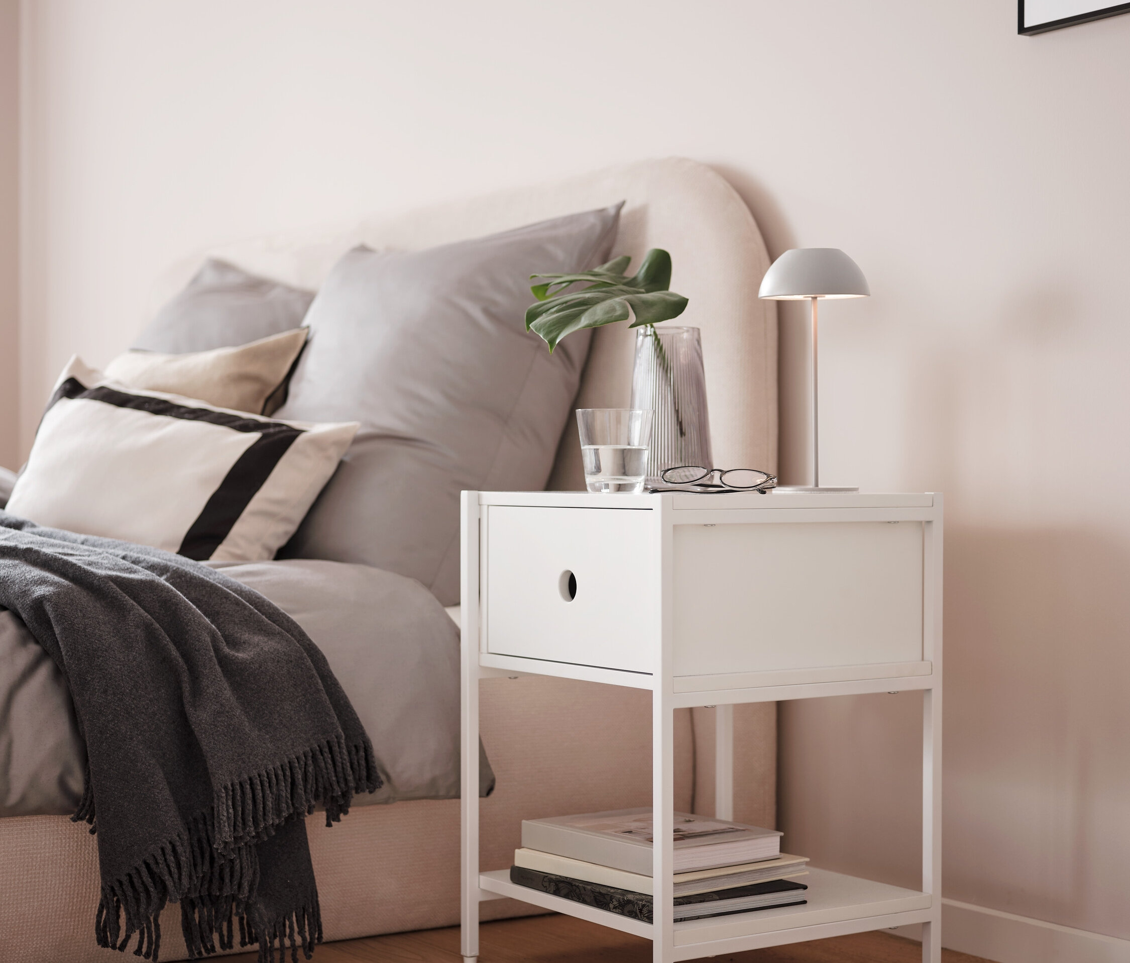 Sivá stolová LED lampa s akumulátorom na bielom nočnom stolíku vedľa postele.