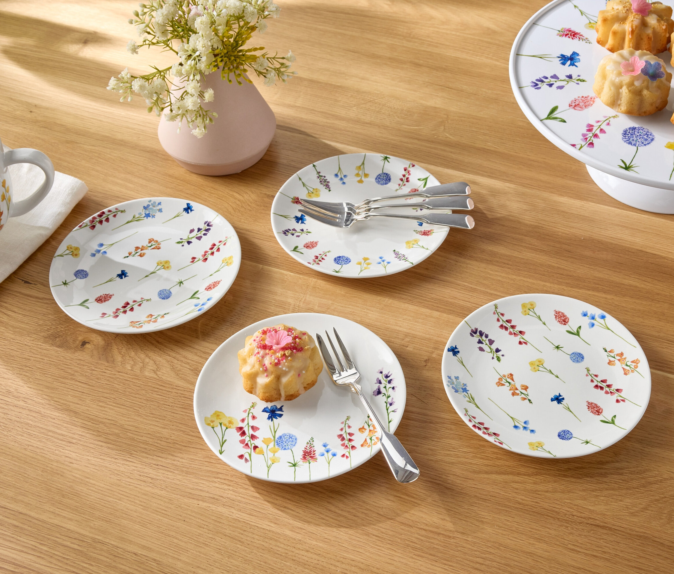 Na drevenom stole je šálka s kávou, váza s kvetmi, taniere a malé koláčiky.