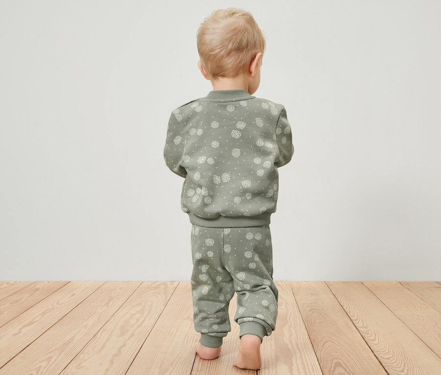 Malé dieťa zozadu kráča po drevenej podlahe a má na sebe zelenú teplákovú súpravu.