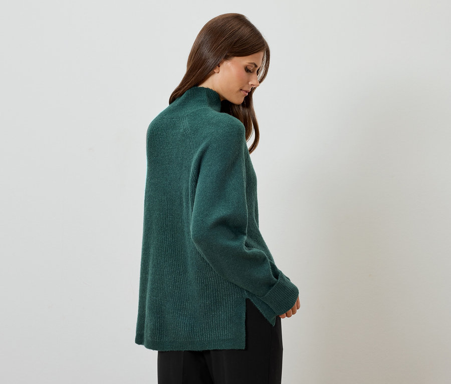 Žena pózuje v zelenom svetri a čiernych nohaviciach.