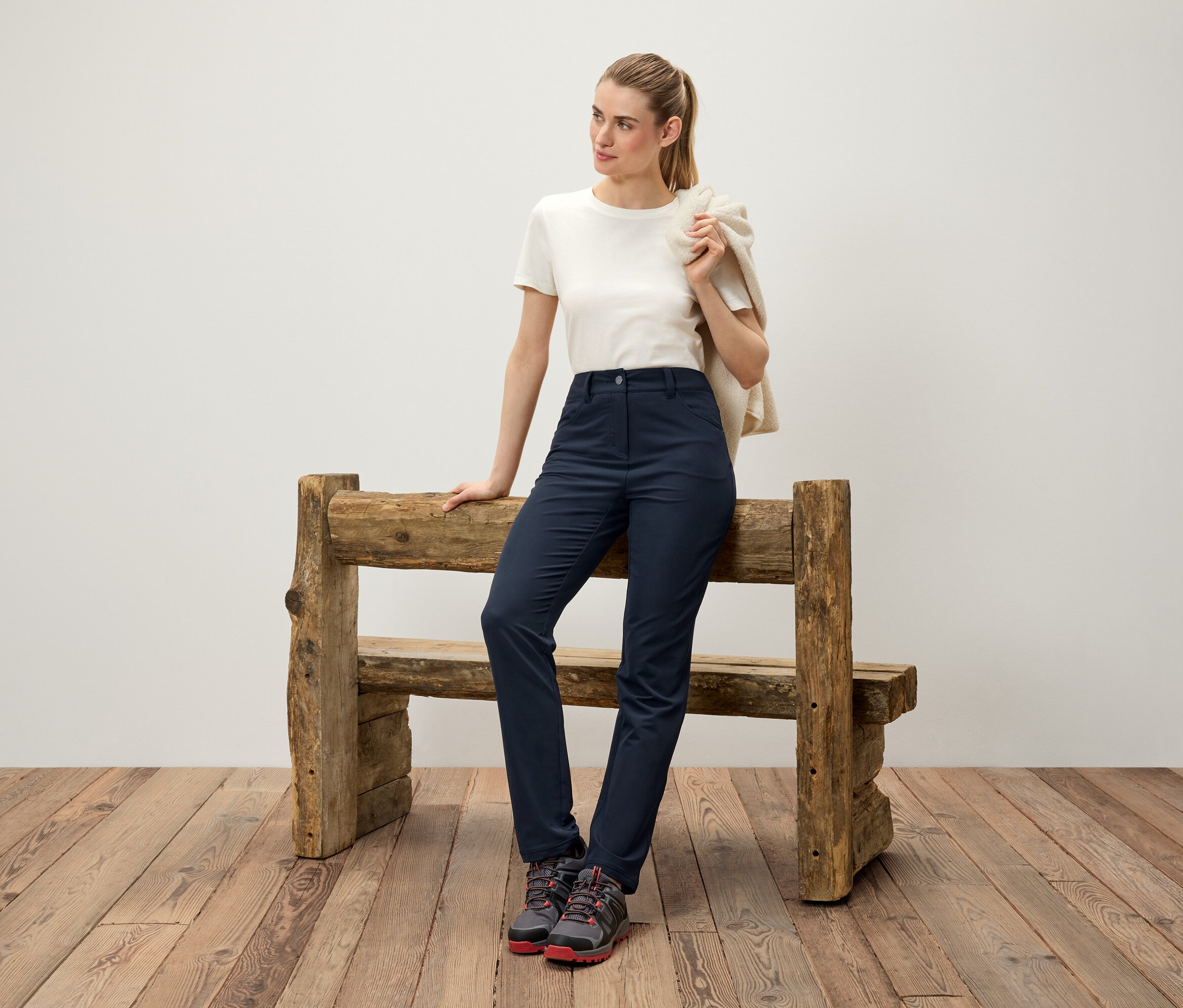 Žena opretá o drevenú lavičku má cez rameno prehodený krémový sveter, na sebe má biele tričko, tmavomodré nohavice a trekové topánky.