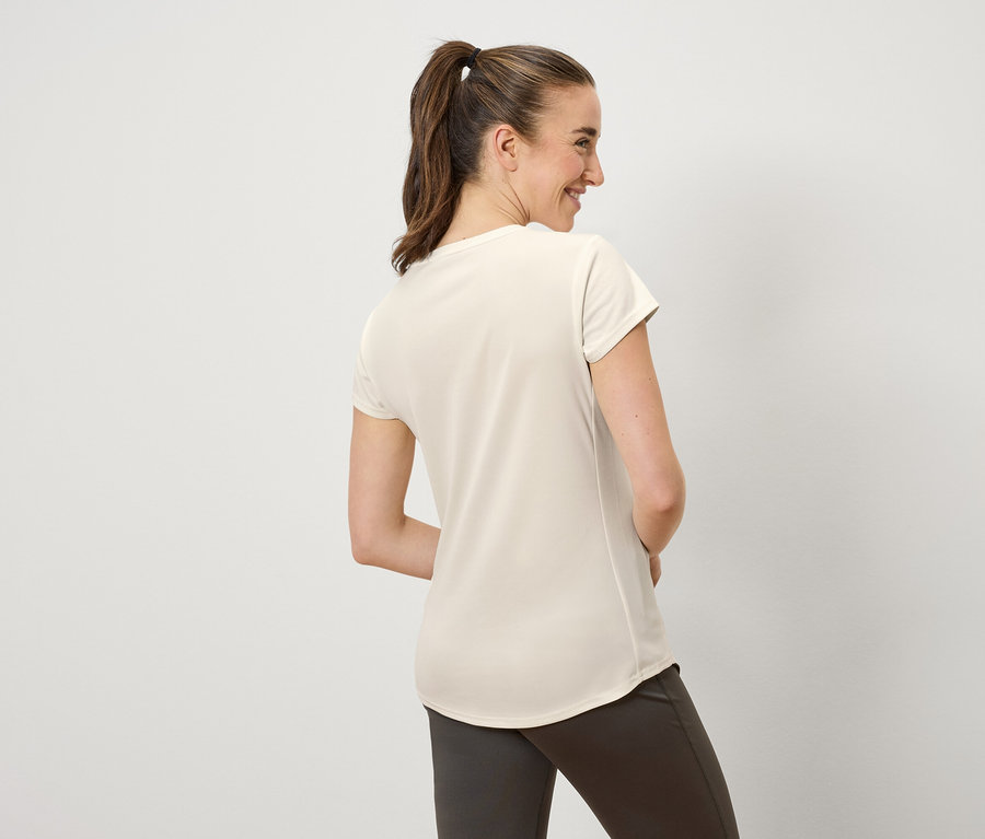 Žena s hnedými vlasmi stiahnutými do copu sa usmieva a pozerá sa doľava. Má na sebe krémové tričko a hnedé nohavice.