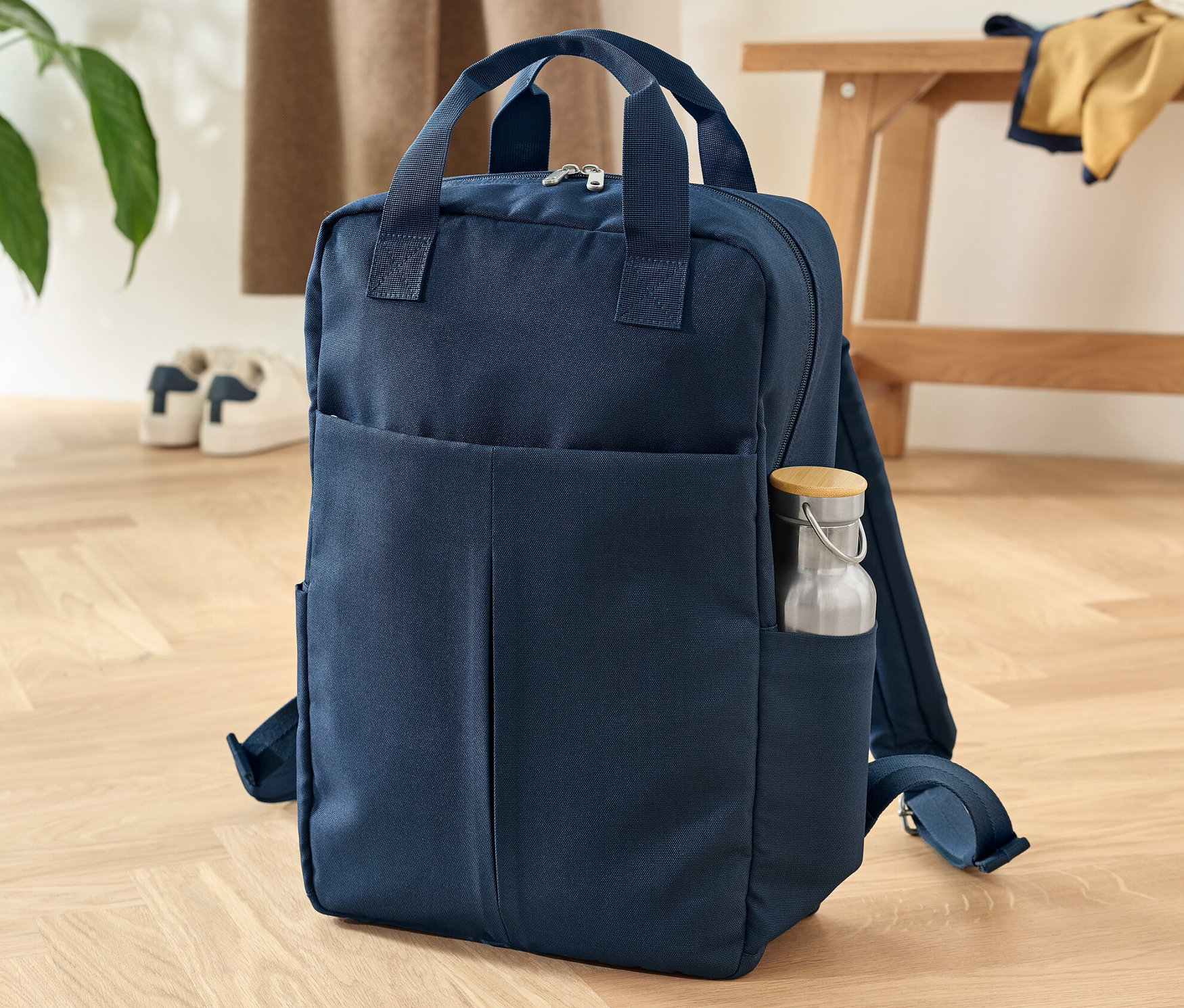 Tmavomodrý bezpečnostný ruksak stojí na drevenej podlahe. V bočnom vrecku je fľaša na vodu.