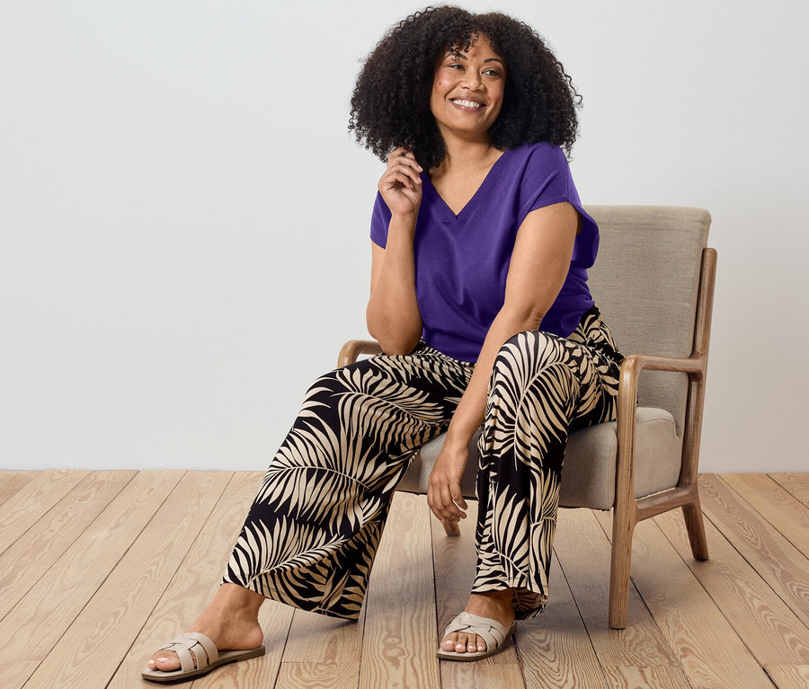 Žena sedí na stoličke a má na sebe fialový pulóver a nohavice v štýle palazzo s potlačou.