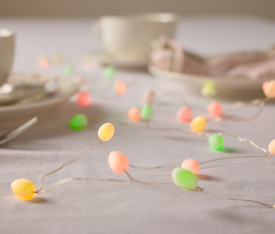 Detailný záber stola s riadom a svetelnou reťazou v tvare veľkonočných vajíčok v pastelových farbách.