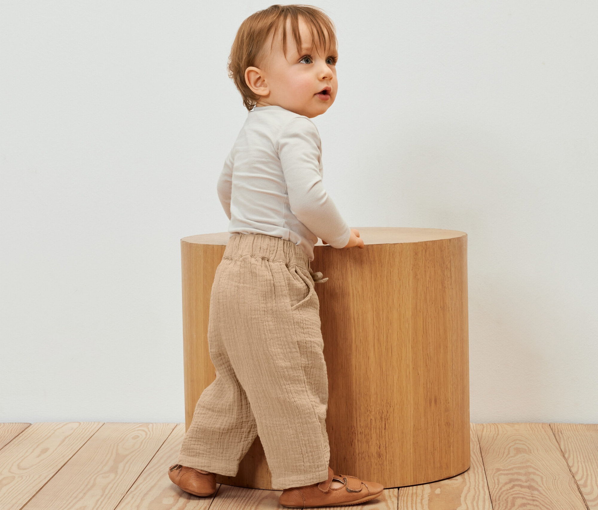 Dieťa stojí vedľa drevenej stoličky a má na sebe detské mušelínové nohavice.