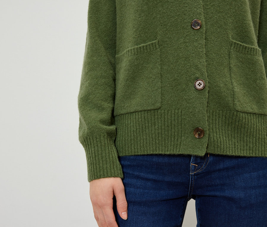 Detail dámskeho olivového svetra s hnedými gombíkmi a vreckami, oblečeného s tmavomodrými džínsami.