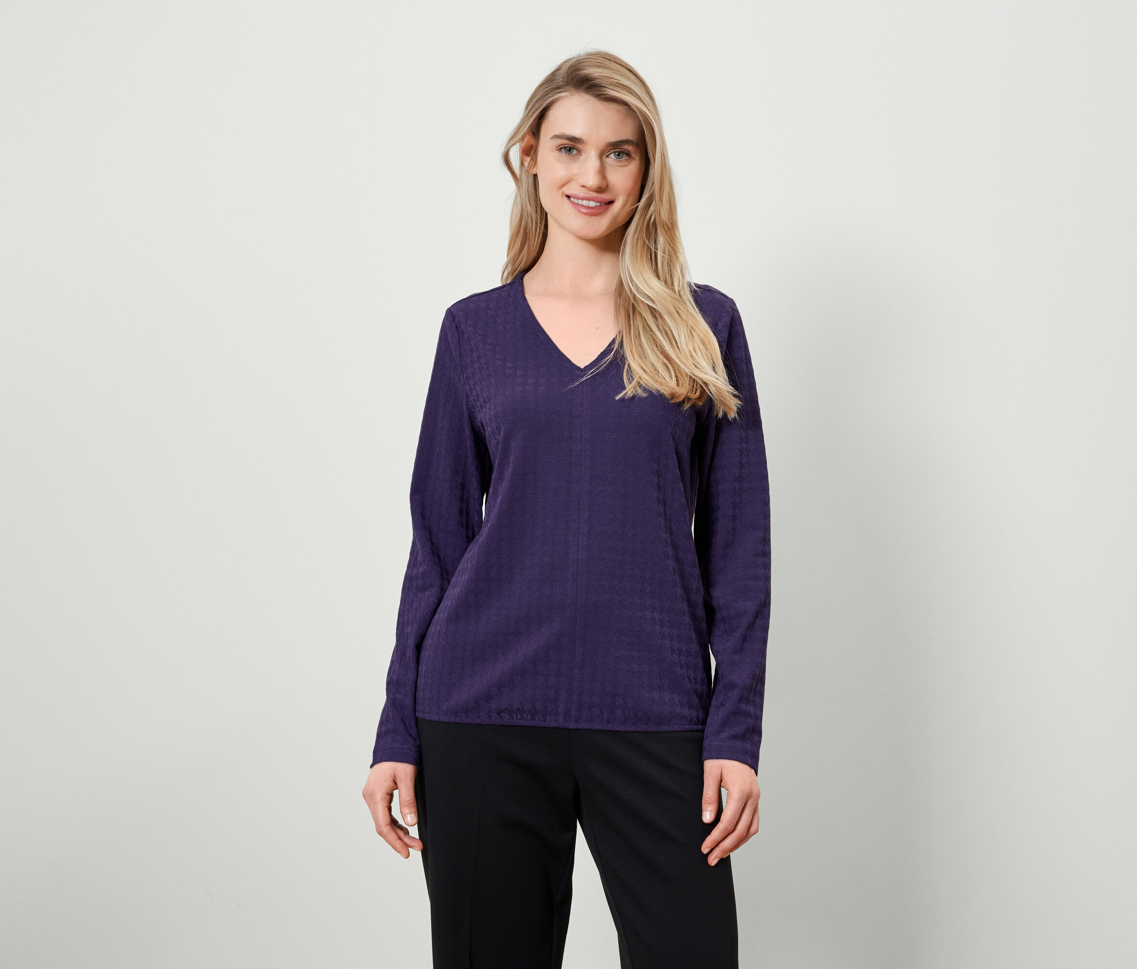 Blondínka s dlhými vlasmi pózuje v purpurovej blúzke s dlhými rukávmi a čiernych nohaviciach.