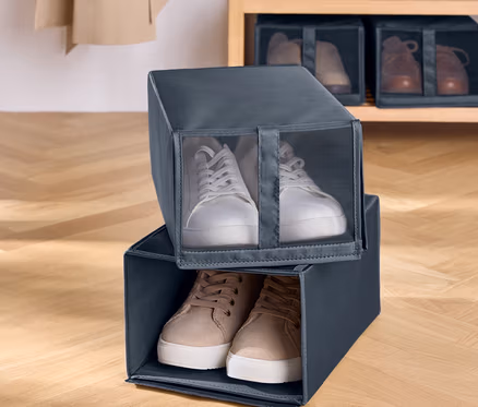 Dva sivé textilné boxy na topánky sú naskladané na seba. V hornom boxe sú biele tenisky a v dolnom hnedé tenisky. V pozadí je drevený regál s topánkami.