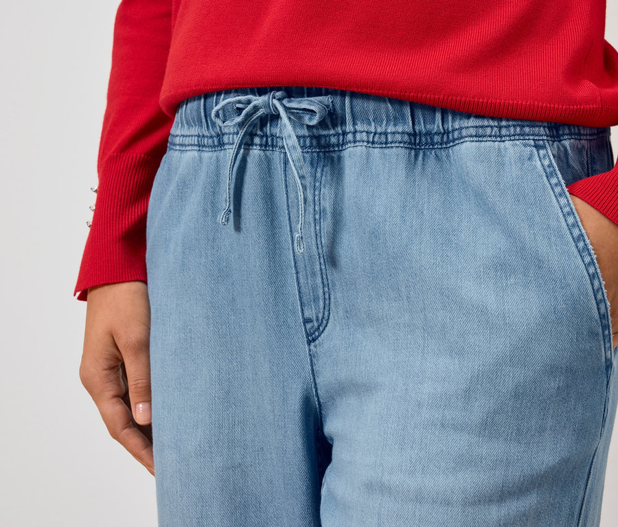 Čiastočný pohľad na osobu v červenom svetri a svetlomodrých džínsoch s rukou vo vrecku.