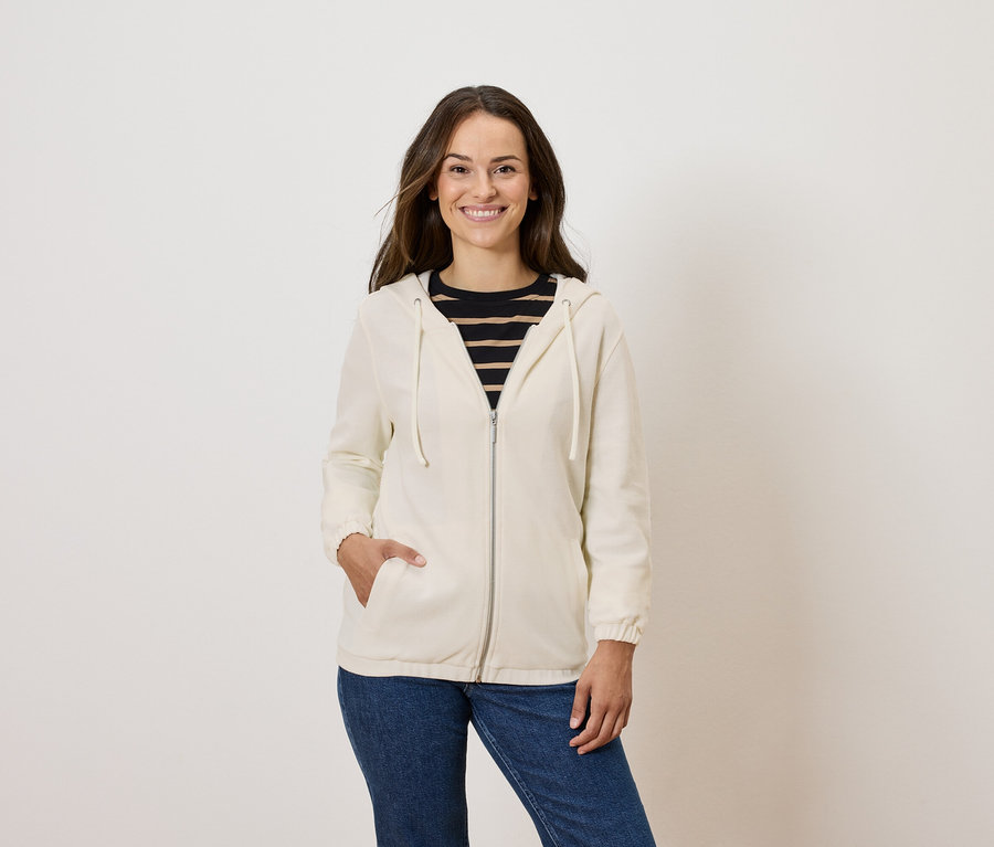 Žena pózuje v krémovej mikine na zips a modrých džínsoch.