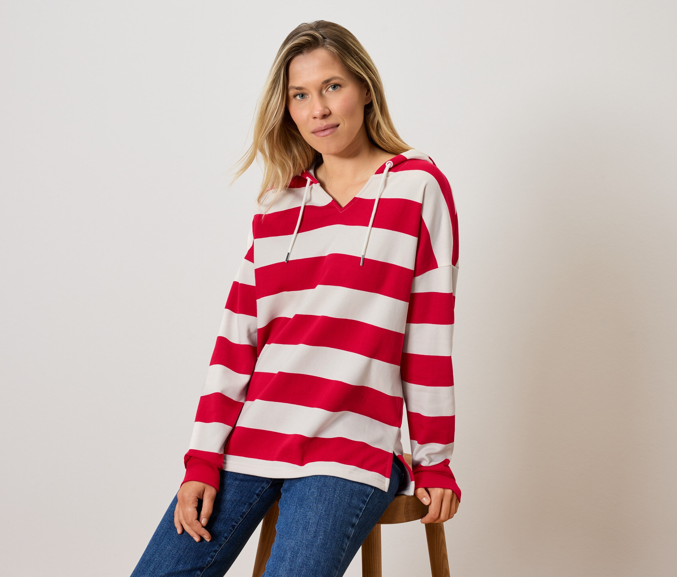 Žena sedí na stoličke a má na sebe pruhované tričko s kapucňou a džínsy.