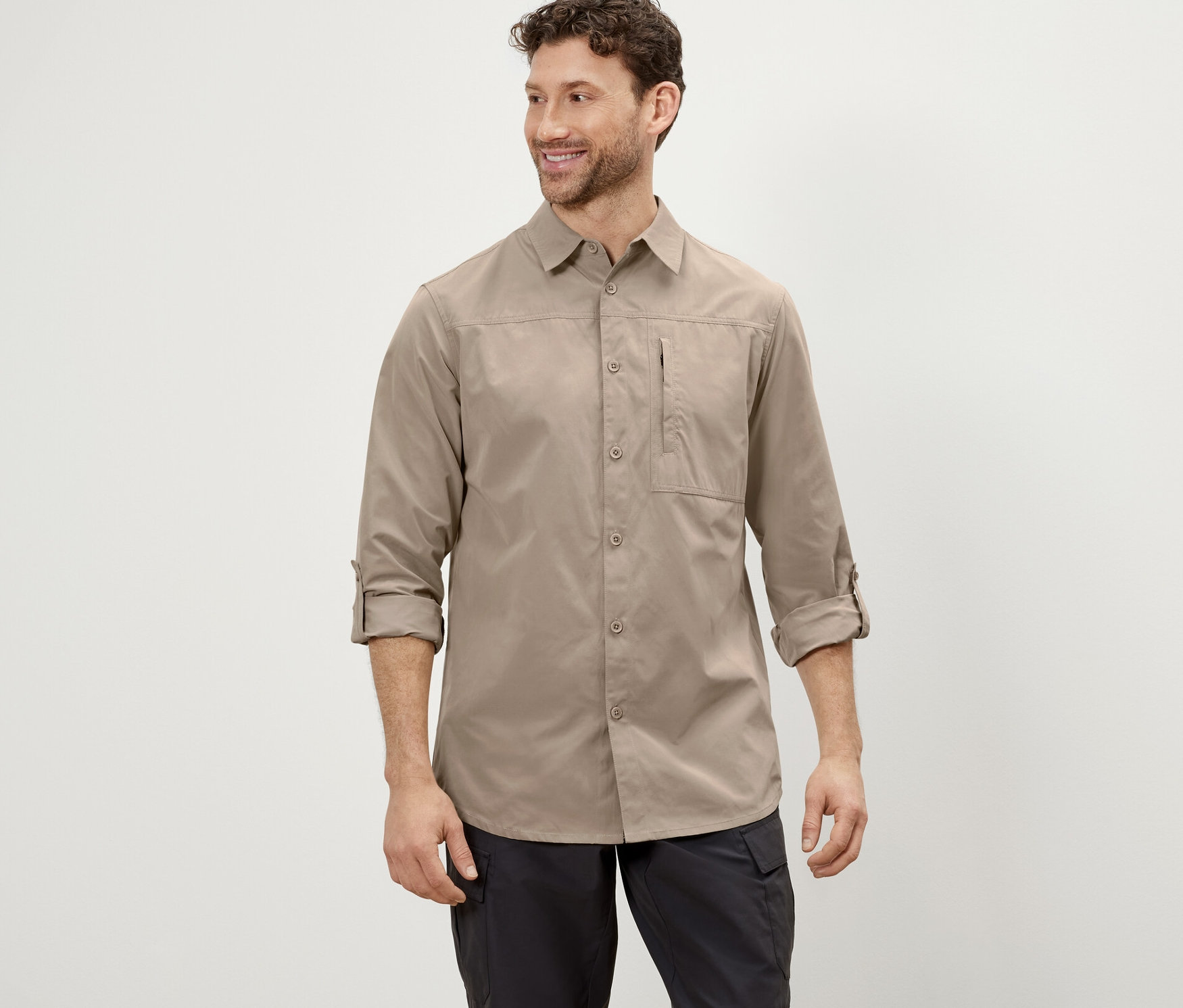 Muž pózuje v béžovej outdoorovej košeli s ochranou proti komárom a čiernych nohaviciach.