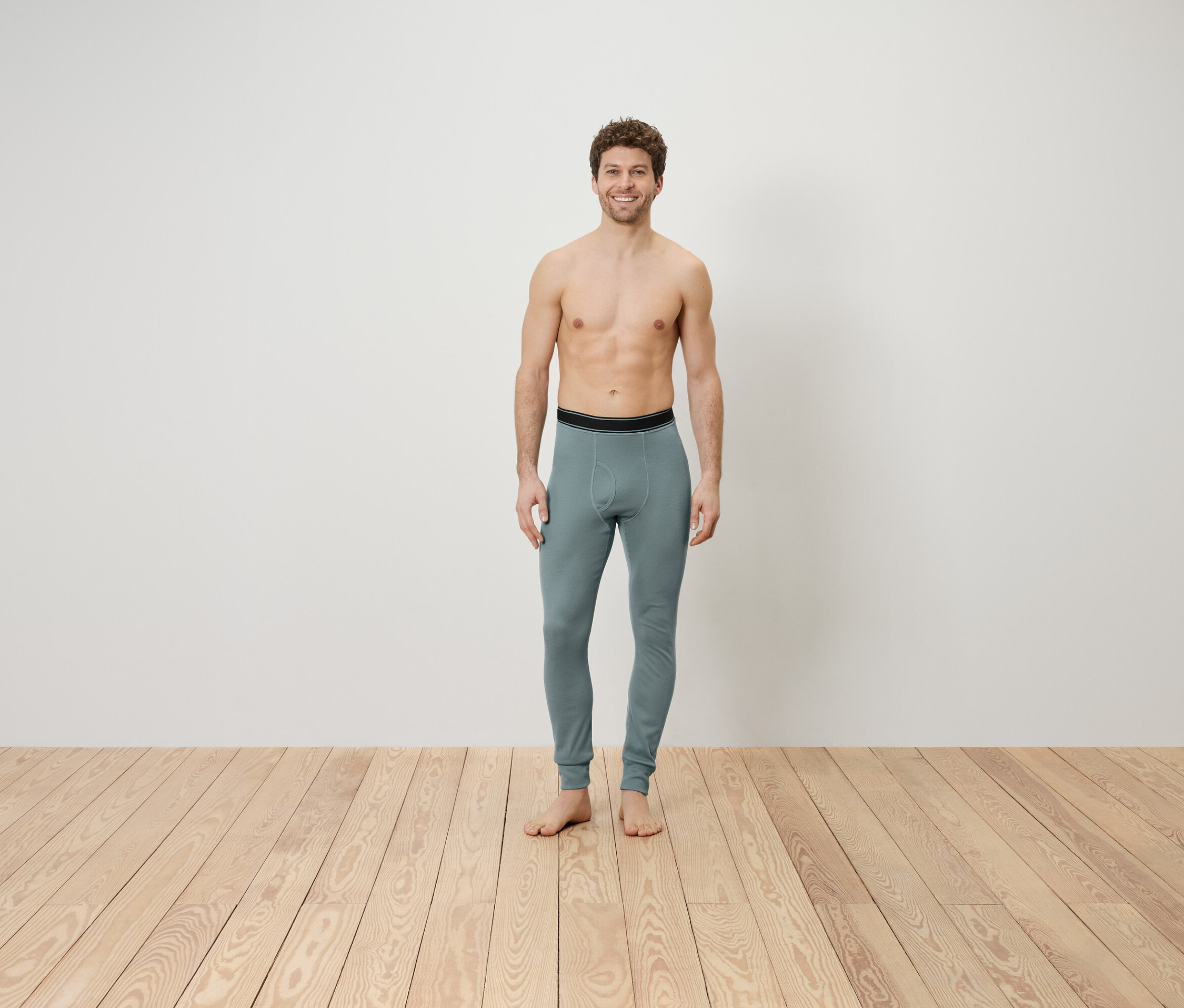 Muž s odhalenou hornou časťou tela stojí na drevenej podlahe a má na sebe sivé legíny.