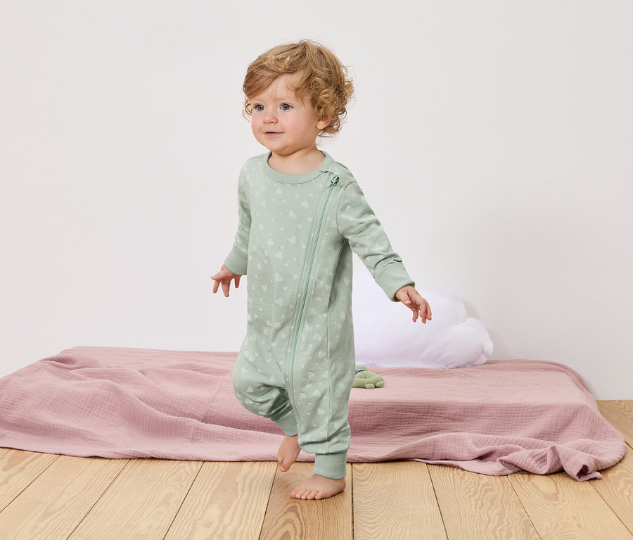 Malé dieťa s kučeravými blond vlasmi kráča bosé po drevenej podlahe. Má na sebe svetlozelené pyžamo so zipsom a vzorom srdca.