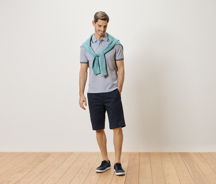 Muž stojí so svetlomodrým svetrom okolo krku, sivou polokošeľou, tmavomodrými šortkami a modrými teniskami.