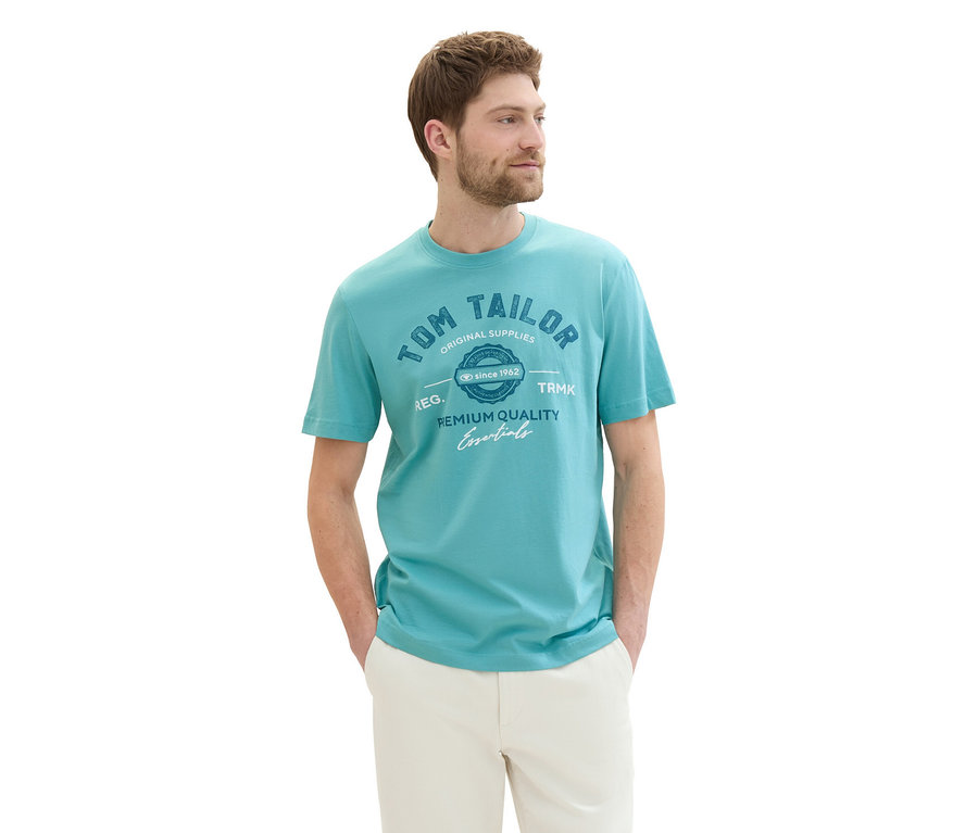 Muž v tričku s logom Tom Tailor, meadow teal a béžových nohaviciach s rukami vo vreckách.