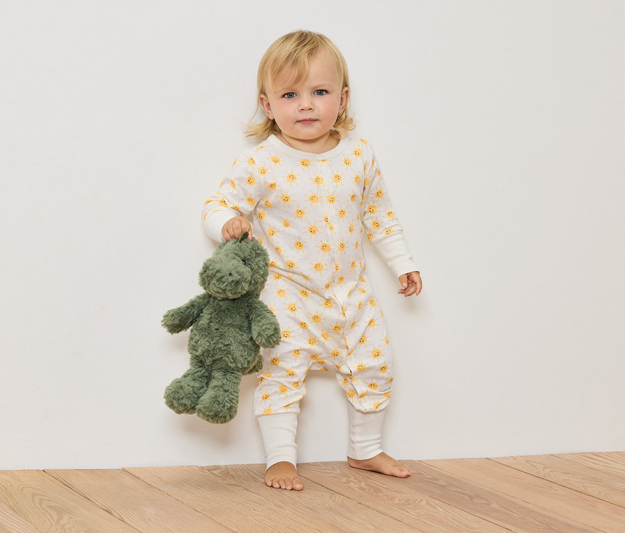 Blond dieťa v overale so vzorom slnka drží zeleného plyšového dinosaura.
