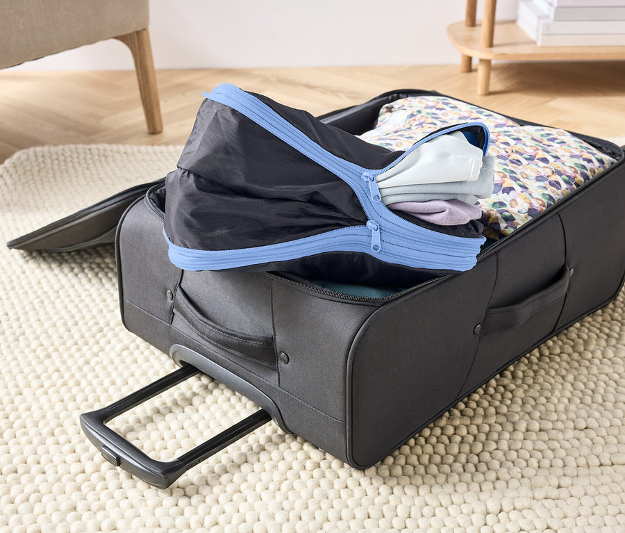 Otvorený, sivý kufor na koberci, vyplnený oblečením a čiernou baliacou kockou s modrým zipsom.