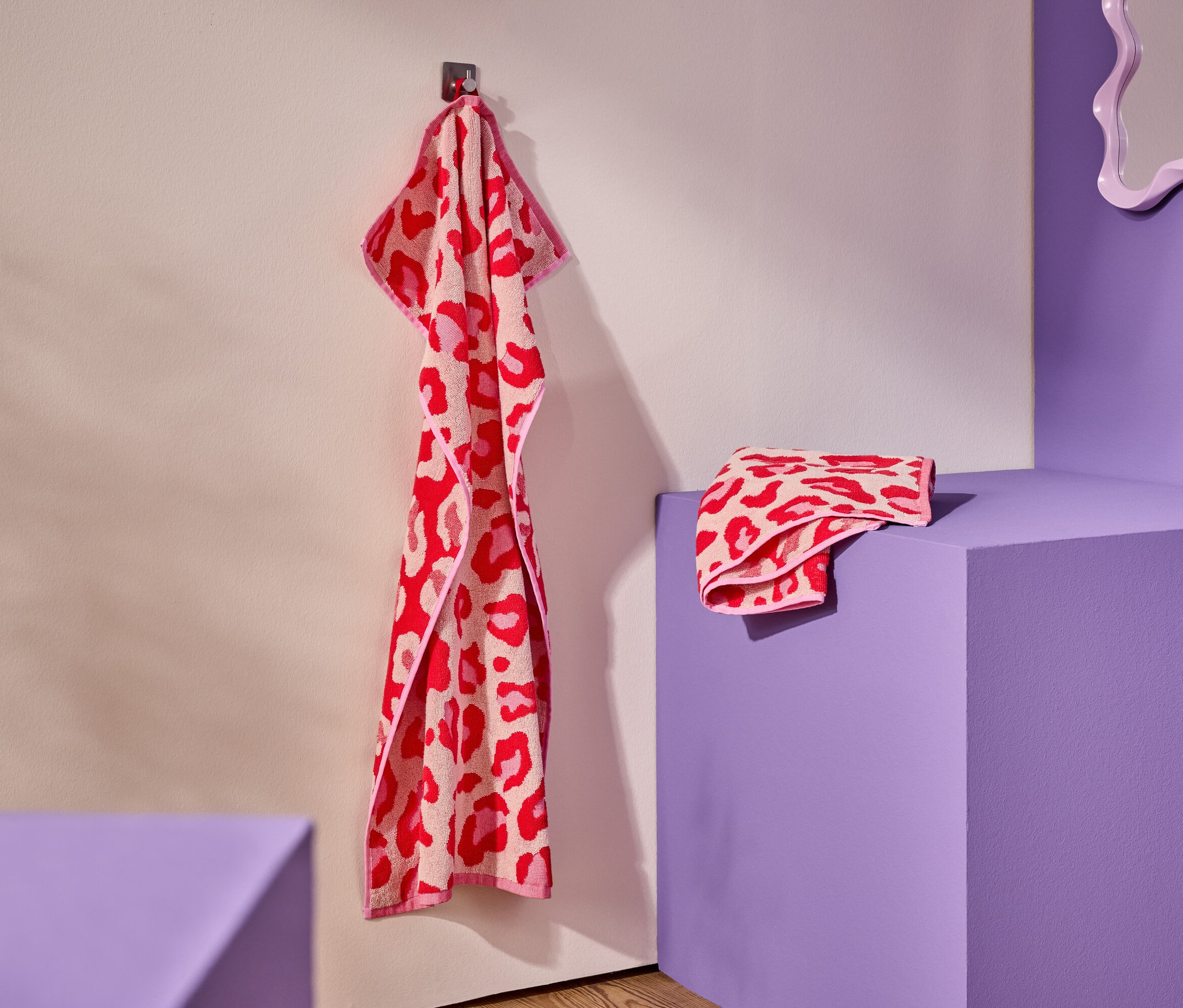 Červeno-ružový uterák s leopardím vzorom visí na háku. Zložený uterák leží na fialovom podstavci.