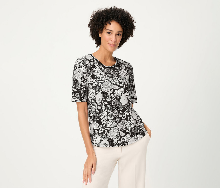 Žena s kučeravými vlasmi má na sebe tričko Olsen s kvetinovým vzorom a svetlé nohavice.